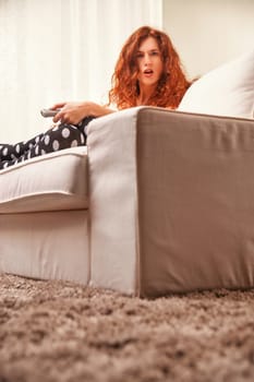 Young redhead woman sitting on a white sofa in her cozy living room, holding a remote control and watching a dull tv program, expressing boredom and frustration
