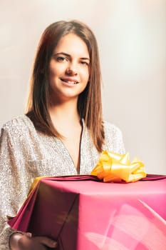 Young smiling woman holding a big pink gift with a yellow bow, looking at camera, posing in studio on white background, expressing happiness and excitement for a special occasion