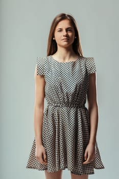 Studio portrait of a serious young woman standing still with long brown hair and a short dress, looking directly at the camera with a neutral expression