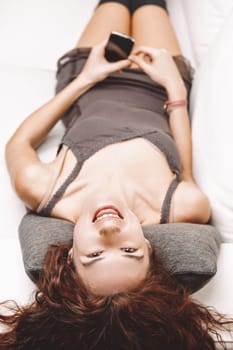 Young woman with long, curly red hair lying upside down on a white sofa, smiling while holding a smartphone, radiating happiness and enjoying a moment of relaxation indoors