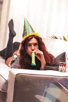 Young woman wearing a colorful party hat lies on a sofa, blowing a party horn and joyfully celebrating her birthday alone at home in a cheerful, festive atmosphere
