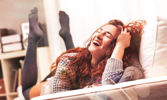 Cheerful young woman with long, red curly hair is lying on a comfortable sofa with her feet up, laughing heartily and enjoying a relaxing moment at home