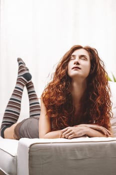Serene young woman with long, curly red hair resting on a comfortable sofa, eyes closed and embracing a peaceful moment of tranquility and relaxation in her cozy living room