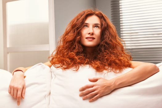 Young woman with long, curly red hair is lying on a white sofa, resting her arms on the back and looking upwards with a thoughtful expression