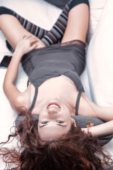 Young woman with long, curly red hair smiling while lying upside down on a white sofa, dressed in a stylish gray dress and playful striped socks, radiating joy and relaxation