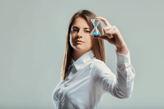 Young businesswoman is holding a small hourglass, looking at the camera with a serious expression, symbolizing the importance of time management in a professional setting