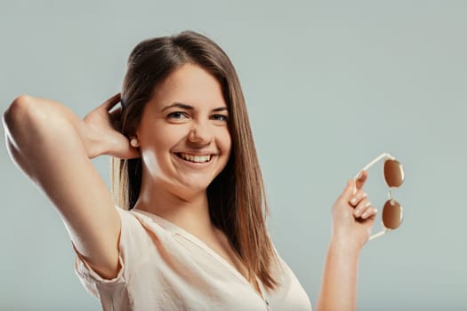 Studio portrait of a cheerful young woman holding sunglasses and running her fingers through her hair, expressing happiness and carefree attitude