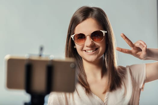 Smiling teenager girl wearing sunglasses is making the peace sign with her fingers while recording a video blog or selfie with her smartphone on a tripod