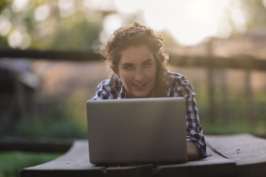 Young woman lying on a wooden table outdoors, engaging with a laptop while looking at the camera with a playful smile, surrounded by the tranquility of a rural setting