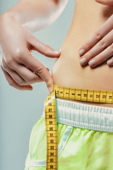 Close-up of a young woman pinching her belly fat while measuring her waist with a yellow measuring tape, expressing concerns about her weight and body image