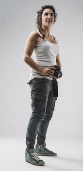 Young female photographer standing in a studio, holding a camera and gazing thoughtfully upwards, embodying creativity and inspiration in her casual tank top and cargo pants