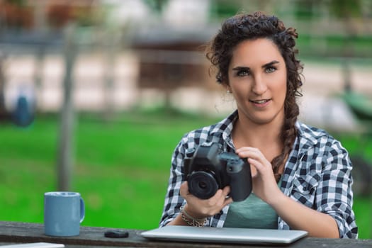 Young female photographer sitting at a table in a park, checking photos on her digital camera while enjoying a cup of coffee and working on her laptop amidst nature's greenery