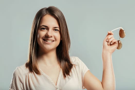 Studio shot featuring a beautiful young woman joyfully smiling at the camera while holding a stylish pair of sunglasses, radiating confidence and happiness against a gray background