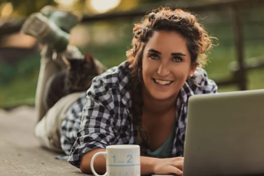 Young woman lying on a wooden deck, working on her laptop while her cat peacefully rests on her legs, soaking up the fresh air and warm sunshine in a serene outdoor setting