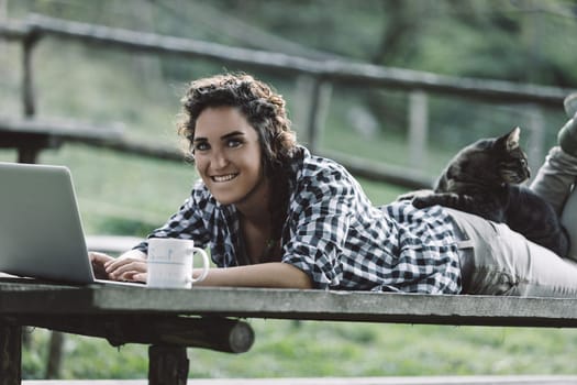Young woman lying on a wooden picnic table, working on her laptop while sipping coffee. A cat comfortably resting on her back adds a cozy touch to the serene outdoor scene