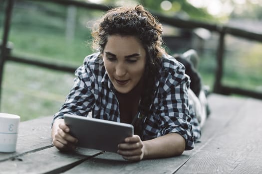 Young woman lying on a wooden deck, wearing a checkered shirt, smiling while using a digital tablet. A mug sits nearby, creating a serene atmosphere on the farm