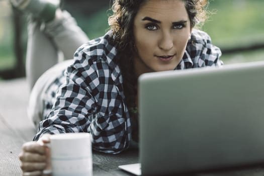 Young woman lying on a wooden table outdoors, working on her laptop while holding a mug of coffee, smiling confidently and looking directly at the camera in a relaxed, casual setting