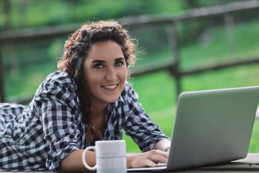 Young woman is lying on her stomach on a picnic table, working on her laptop with a mug next to her, enjoying the fresh air and greenery of a park