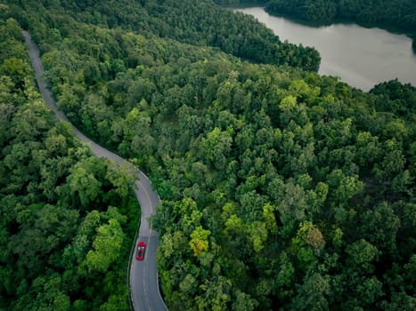 Aerial top view of a red car driving on highway road in green forest. Sustainable transport. Electric vehicle driving on asphalt road through lush green forest. Zero emission car. Green mobility.
