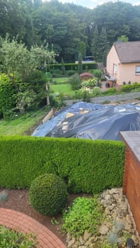 A construction site in a suburban neighborhood, featuring an excavated area covered with a tarp, a wooden shed, and residential houses in the background.