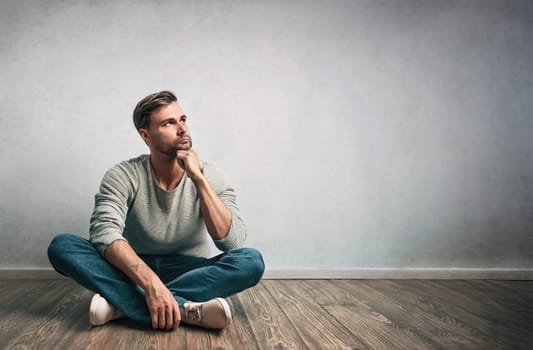 Relaxed man sitting on the floor with hand under chin looks pensive aside on blank background with copy space. Thoughtful male seeking for new ideas