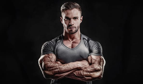 Brave man athlete portrait standing with arms crossed, looking confident over dark background. Strong sportsman with muscular arms. Bodybuilder and workout concept