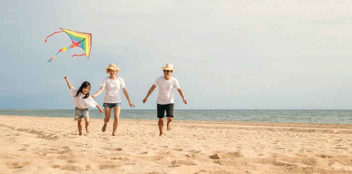 Asian family is flying a kite high in the sky while running on the beach. Happy parents mother and father with their child playing with kite, family is enjoying their time together, summer day