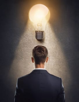 Closeup rear view thoughtful businessman inside a dark room with a glowing lightbulb above head. Bulb as symbol for genius idea, inspiration and creativity