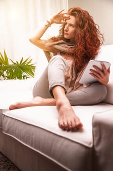 Young woman with long red hair relaxing on a white sofa, holding a tablet in one hand while shielding her eyes from sunlight streaming through the window