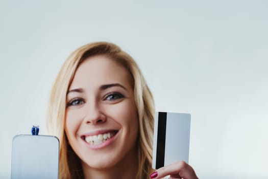 Young adult woman holding a smartphone with a small blue gift box on top and a credit card, smiling and looking at camera, on a white background, suggesting online shopping for gifts