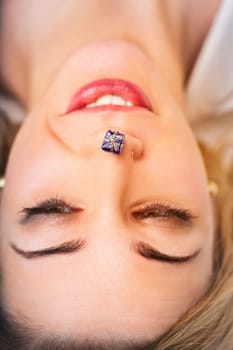 Young woman lying down, smiling brightly with a tiny wrapped gift balanced playfully on her nose, embodying joy, surprise, and a carefree spirit in a festive moment