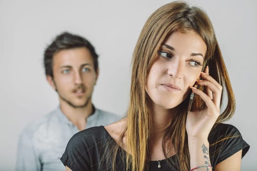 Young woman talking on a mobile phone, deliberately ignoring her jealous boyfriend in the background, creating a tense atmosphere filled with unspoken emotions and conflict