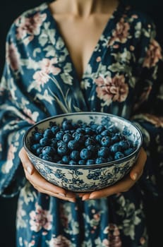 A woman in a floral robe holds a large bowl filled with fresh blueberries, capturing a moment of healthy indulgence.