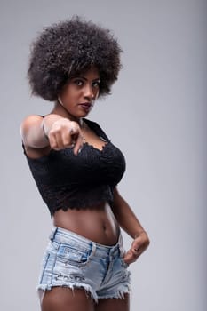 Young woman with afro hair pointing her finger at the viewer, displaying a serious expression. Dressed in jeans shorts and a black top, exuding confidence and assertiveness