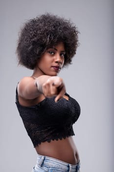 Young woman with a stylish afro hairstyle, dressed in a black top and jeans, confidently pointing her finger with a determined expression in a modern studio setting