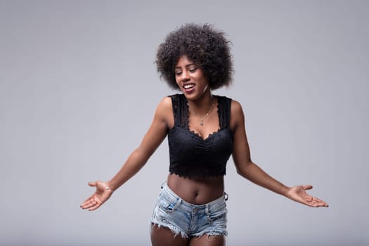 Young woman with an afro hairstyle is shrugging her shoulders with open arms and a confused expression, looking down and to the side against a gray background