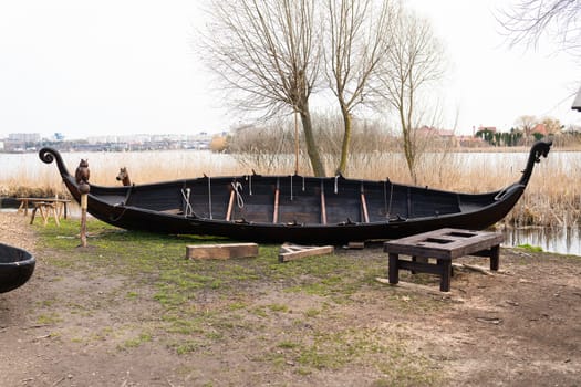 A traditional Viking boat on a grassy shore near the water, with wooden benches nearby and reeds in the background.