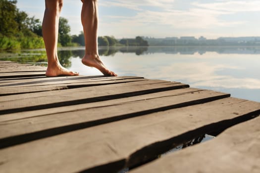 Close-up of bare feet on a wooden dock with a serene lake and trees in the background under a bright sky.