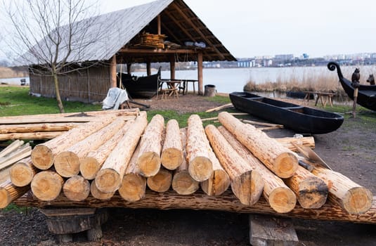 A pile of timber logs near a lakeside workshop, with wooden boats, a barn, and water visible in the background.