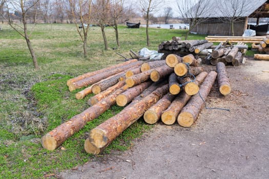 A neat pile of freshly cut logs stacked in a rural yard, surrounded by trees and green grass under a cloudy sky.