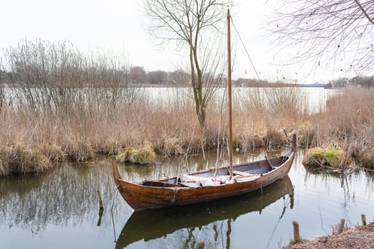 A wooden Viking-style boat resting on a still lake surrounded by reeds and bare trees under a cloudy sky.