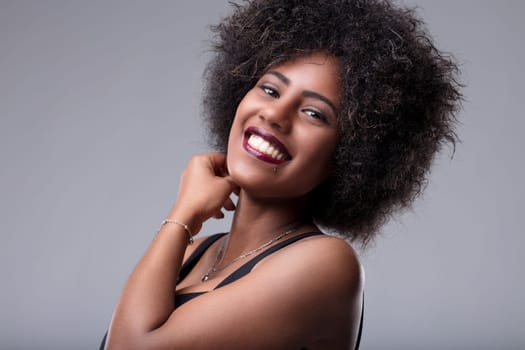 Studio portrait of a cheerful young woman with voluminous afro hair, touching her neck while flashing a captivating smile, exuding confidence and positive vibes