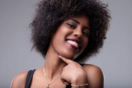 Studio portrait of a cheerful young woman with afro hair, smiling and touching her chin with her finger, expressing confidence and happiness