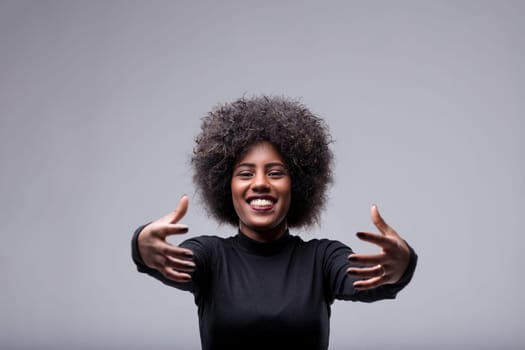 Studio shot of a young woman with afro hair, wearing a black turtleneck, opening her arms for a hug, smiling and looking at the camera, expressing joy and affection