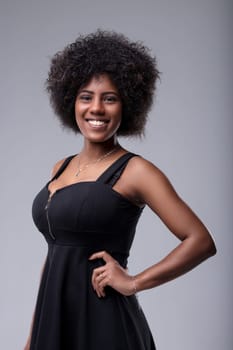 Studio portrait of a cheerful young black woman with curly hair, wearing a black dress, smiling confidently while posing with her hand on her hip against a gray background