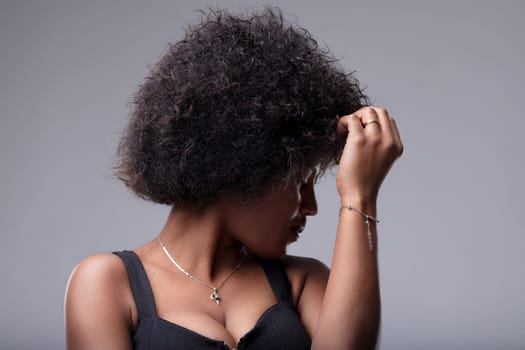 Studio portrait of a young black woman touching her afro hair while looking down, expressing shyness and introspection on a gray background
