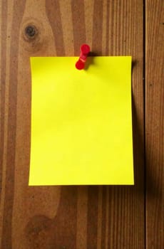 A bright yellow sticky note is pinned to a rustic wooden surface. The note is blank, providing a space for reminders or messages. The wooden background adds a warm, natural feel to the setting.