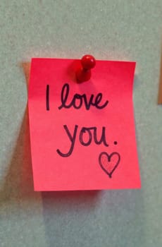 A bright pink sticky note is pinned to a gray wall, showcasing a handwritten message that reads I love you. accompanied by a small heart illustration, creating a warm, affectionate atmosphere.