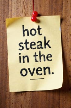 A yellow sticky note hangs on a wooden surface, announcing that a hot steak is in the oven. The note serves as a reminder while evoking a casual cooking atmosphere.