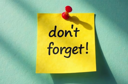 A bright yellow sticky note displays the message don't forget against a teal background. The note is secured with a red pushpin, casting a soft shadow in the sunlight.